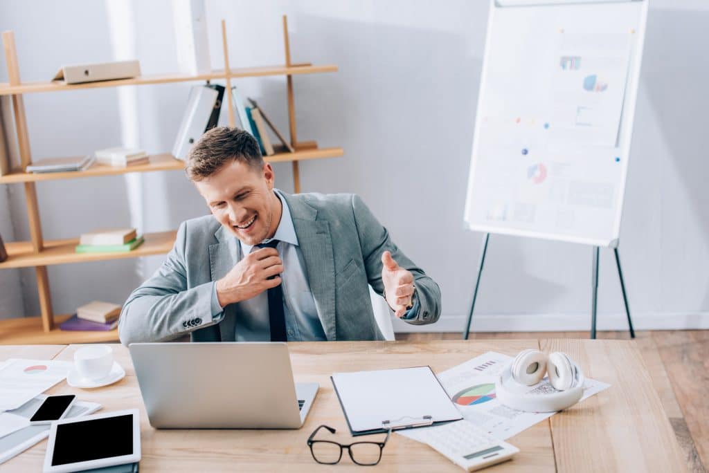 Smiling businessman having video call on laptop near devices, papers and headphones in office - Big Easy SEO